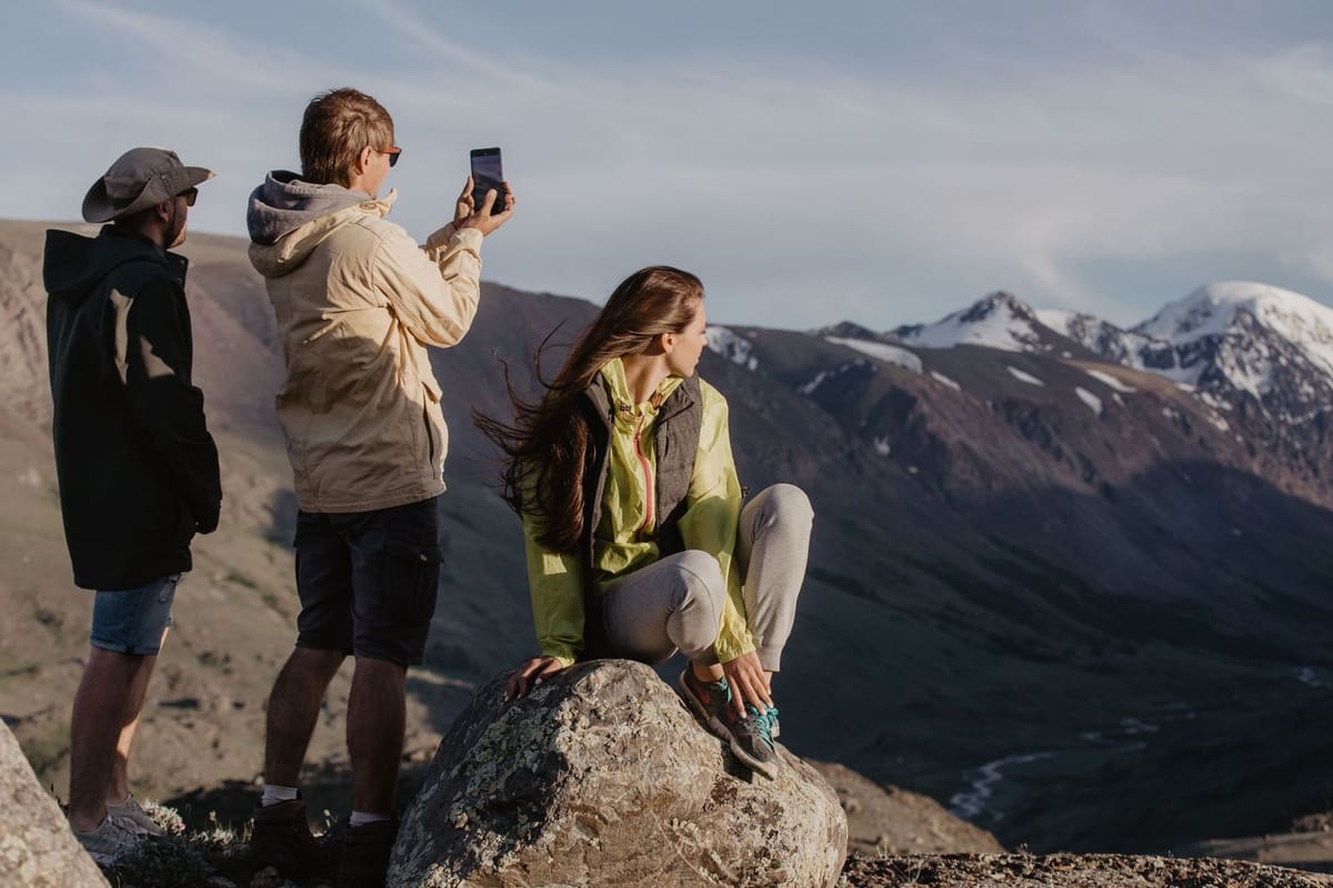 Three hikers on a rock at a summit, one taking a photo of the view